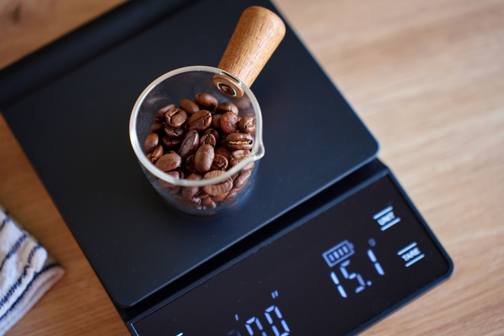 Roasted coffee beans in a glass scoop on a black digital scale on a wooden table. Concept of coffee preparation, measuring beans and drip coffee brewing