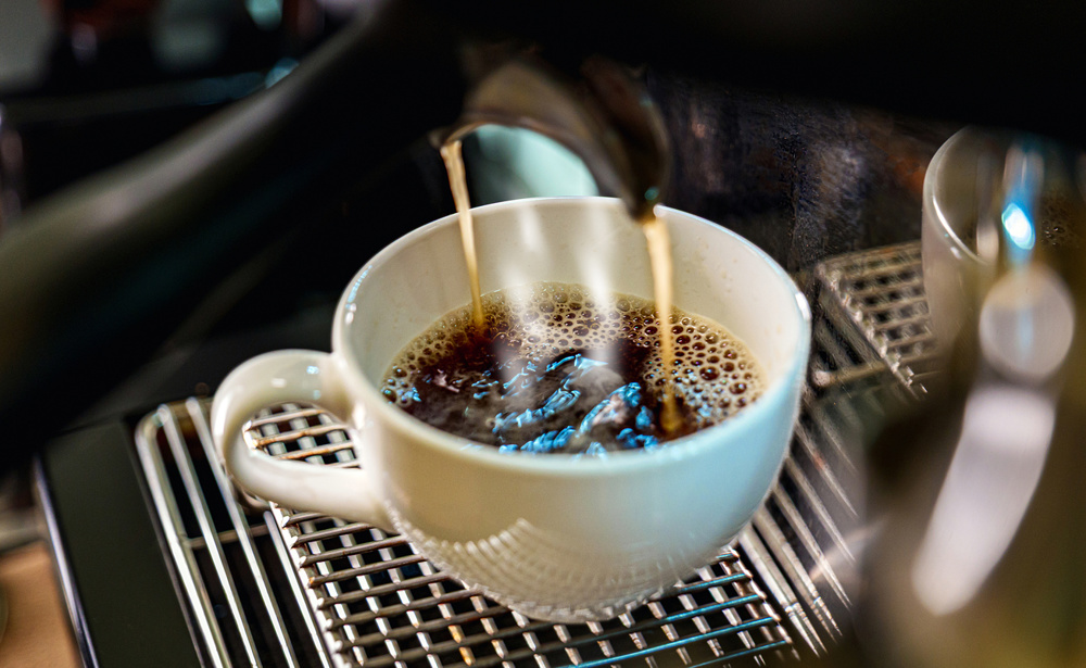 Close-up of rich dark coffee being poured into a white ceramic cup on a professional espresso machine drip tray, capturing the perfect brewing moment for café and lifestyle concepts