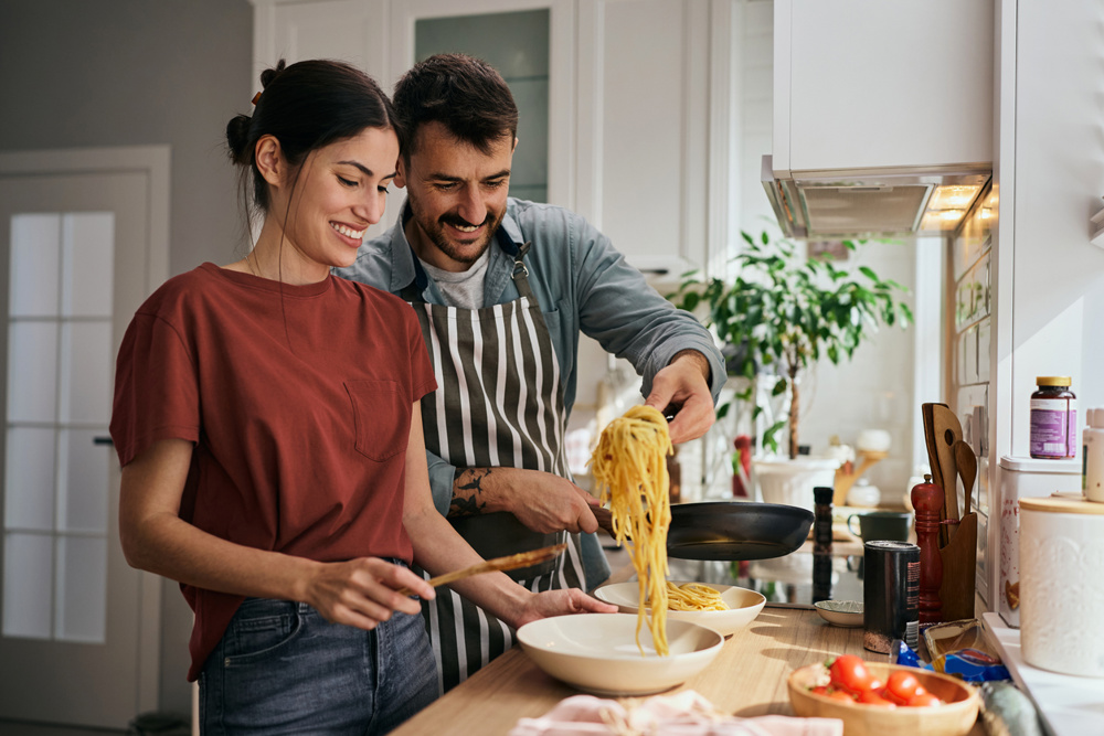 Happy man serving his girlfriend spaghetti while cooking in the kitchen