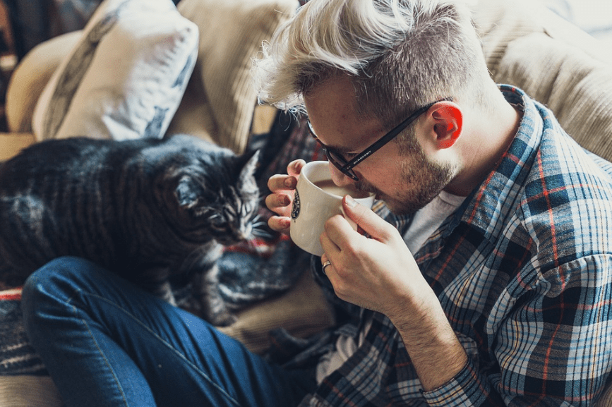 a man on a sofa, sipping a cup of coffee, and a cat