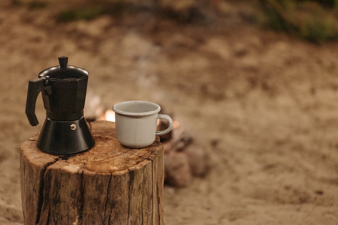 A portable espresso maker on a wooden log with a ceramic cup beside it
